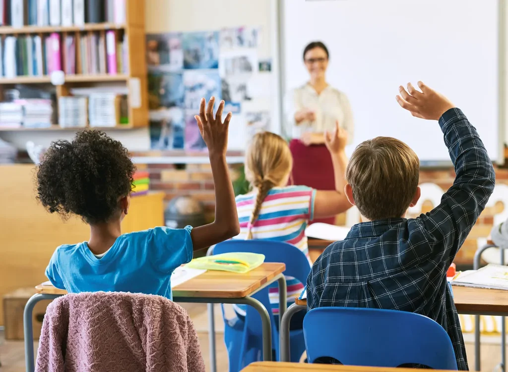 A photograph from the rear of the classroom showing elementary school children from behind raising their hands, while their teach calls on them from the front of the class.