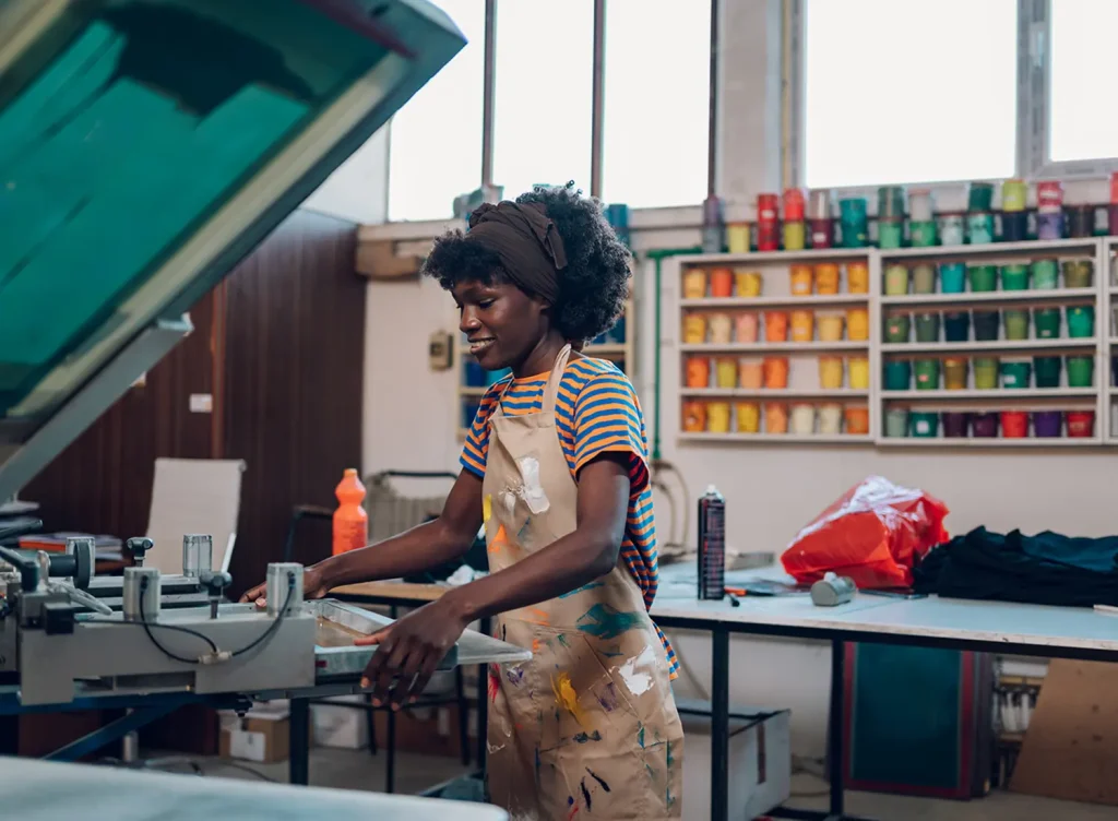 A photograph of a young Black woman using the screen printing setup in a print shop.